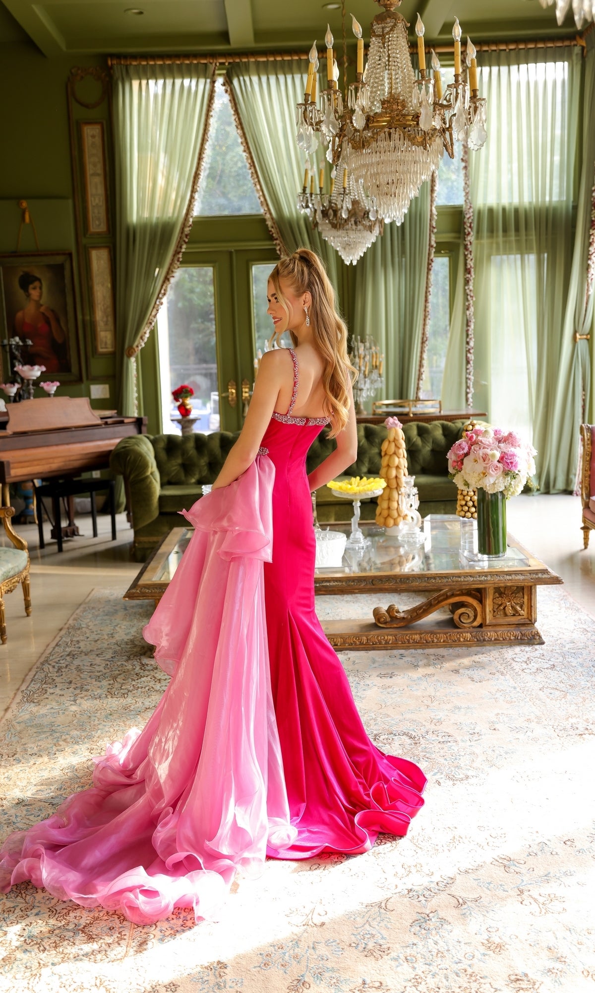 A woman wearing the Long Prom Dress 39556 by Ava Presley stands on a patterned rug in an elegant, sunlit room with green walls, chandeliers, and large windows.